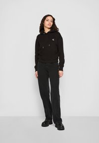 Μαύρη κουκούλα με λογότυπο, πλεκτές μανσέτες και κορδόνι. Συνδυασμένη με φαρδιά μαύρα τζιν και χοντρά μαύρα παπούτσια, μπροστά από έναν απλό φόντο.