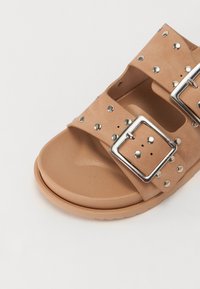Sandálias cor de tan com duas tiras largas, detalhes em fivela prateada e decoração com tachas, apresentando uma palmilha texturizada e sola em borracha.