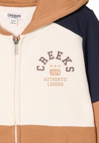 Kapuzen-Zip-Jacke in den Farben Creme, Braun und Marineblau. Material aus Baumwollmix mit Logoprint und gerippten Akzenten an den Ärmeln.