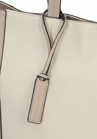 Bolso de cuero beige claro con una superficie texturizada, que presenta una forma curva, bordes negros y una etiqueta de marca adjunta al asa.