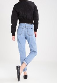 Person klädd i en svart kort jacka, ljusblå jeans uppvikta, och svarta spetsiga ballerinaskor, stående med ett ben korsat bakom det andra.