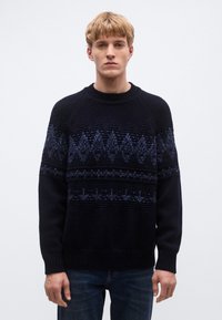 Tengerészkék kötött pulóver különböző kék árnyalatú geometriai mintákkal, bordázott kerek nyakkal és mandzsettákkal, kék farmernadrággal párosítva.