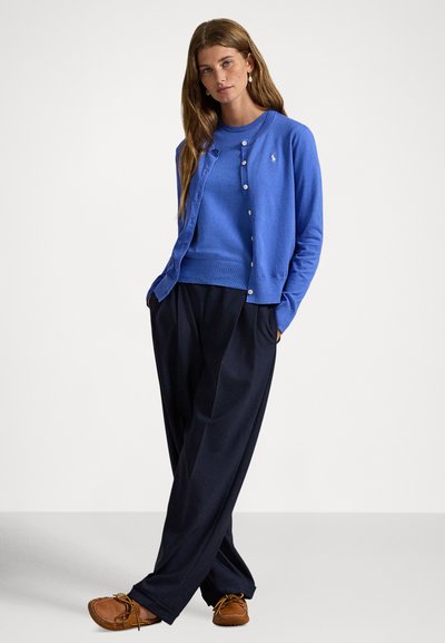 Ανοιχτό μπλε ζακέτα πάνω από το αντίστοιχο πουλόβερ, και τα δύο σε μαλακό πλεκτό ύφασμα. Σκούρο παντελόνι με άνετη εφαρμογή, συμπληρωμένο με καφέ μοκασίνια. Απλά κουμπιά ως διακοσμητικά στοιχεία.