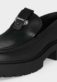 Μαύρο δερμάτινο loafer με χοντρή σόλα και ασημένια αγκράφα μπροστά, με ανάγλυφο το όνομα της μάρκας στην μεταλλική αγκράφα.