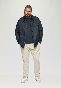 Granatowa koszula z guzikami noszona na ciemnej koszulce, w zestawie z jasnobeżowymi spodniami i białymi trampkami. Krótkie zarost, włosy związane do tyłu.