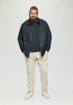 Marineblauw button-up shirt over een donker t-shirt, gecombineerd met lichtbeige broek en witte sneakers. Korte baard, haar teruggebonden.