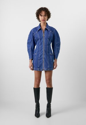 Denim mini haljina s pripijenim siluetom, napuhanim rukavima, prednjim zatvaračem i bočnim džepovima u srednje plavoj boji. Crne čizme do koljena.