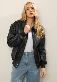 Giacca bomber nera in ecopelle con chiusura a zip, polsini a costine e tasche laterali, indossata sopra una maglietta bianca e jeans blu a vita alta.