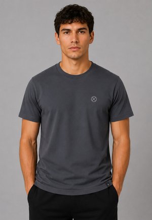 Jeune homme aux cheveux bouclés et foncés portant un t-shirt uni gris foncé à manches courtes avec un petit logo cercle et croix sur la poitrine.