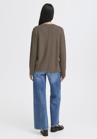 Pull en tricot brun à coupe décontractée, avec des épaules tombantes et une couture au dos, associé à un jean large bleu et des chaussures noires.