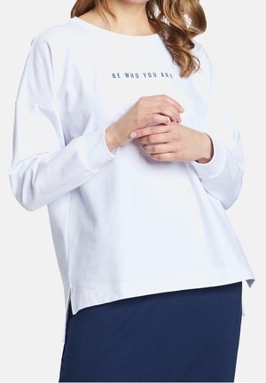 Kobieta w białej koszuli z długim rękawem z napisem „BĄDŹ SOBĄ” i granatową spódnicą, ręce delikatnie splecione z przodu.