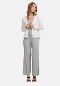 Lichte witte cardigan over een grijze gebreide top en wijde broek, met ribbelstructuur en zijsplitten, gecombineerd met zwarte sandalen.