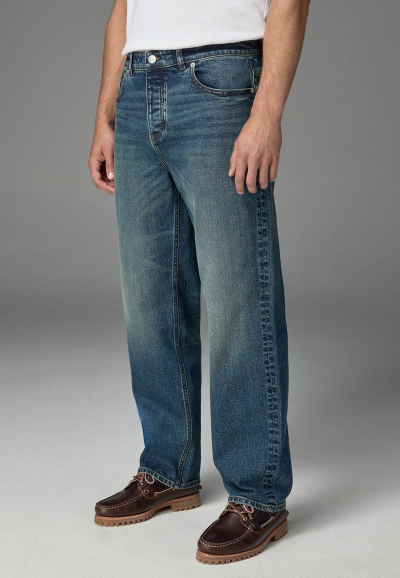 Blugi din denim albastru, cu un finisaj uzat, având cinci buzunare, închidere cu nasture și fermoar, și detalii cusute pe cusături.