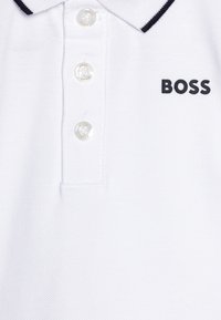 Bela polo majica z teksturirano tkanino, tremi prozornimi gumbi in kontrastnim črnim ovratnikom. Vsebuje logotip "BOSS" v črni barvi.