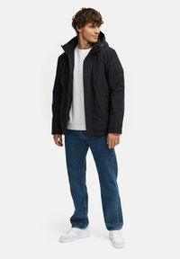 Zwarte buitensjack met een rits aan de voorkant, capuchon en zijzakken, gedragen over een wit T-shirt met ronde hals en blauwe jeans. Witte sneakers.