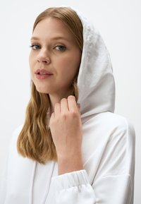 Biała bluza z kapturem, z regulowanym sznurkiem, o miękkiej teksturze i luźnym kroju. Modelka ma długie, falujące włosy i dotyka kaptura.