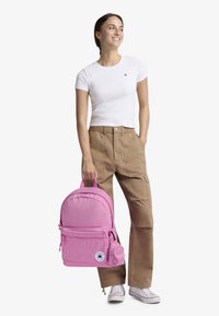 Roosa seljakott, mis on valmistatud siledast kangast, esiküljel tasku, logoga plaastri ja polsterdatud õlarihmadega. Kandmisel beežid cargo püksid ja valge T-särk.