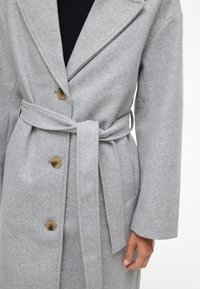 Nærbilde av en person som har på seg en lys grå beltedress med store knapper over en svart topp, og viser en hånd ned langs siden.