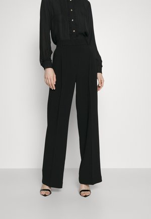 Pantaloni neri a gamba larga con pinces, abbinati a una blouse nera a maniche lunghe con leggere arricciature e bottoni dorati. Tessuto morbido.