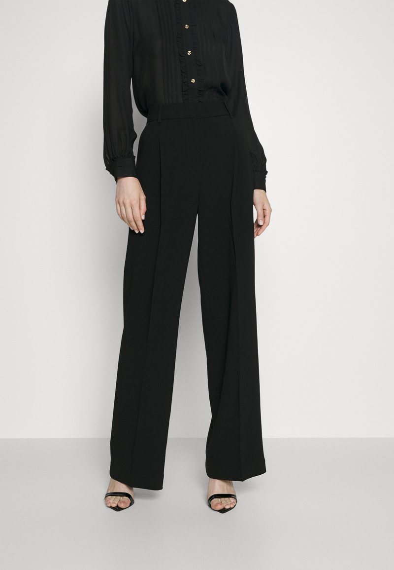 Pantalons noirs à jambes larges avec plis, associés à un blouse noire à manches longues comportant des volants subtils et des boutons dorés. Texture lisse.