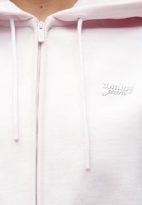 Svetloružová mikina na zips s jemnou textúrou, šnúrkami a vyšívaným logom "tommy jeans" na ľavej strane hrudníka.