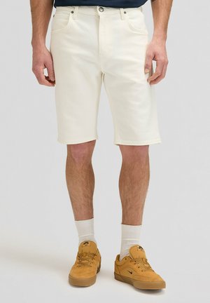 Mann trägt cremefarbene Shorts bis zum Knie, weiße Sportsocken und senfgelbe Nike-Sneaker, steht vor einem einfarbigen Hintergrund.