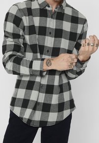 Camisa de cuadros en negro y gris, con botones y mangas largas, que presenta un dobladillo redondeado y botones negros. Tejido texturizado con un patrón de cuadros visible.