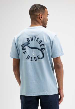 Mann mit kurzen Haaren trägt ein hellblaues T-Shirt mit schwarzer Grafik und dem Schriftzug „Butcher of Blue“ auf dem Rücken, steht vor einem einfarbigen Hintergrund.