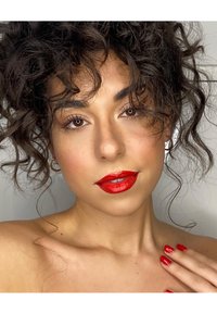 Porträtt av en person med lockigt mörkt hår, röd glansig läppstift och matchande röd nagellack. Subtil glöd på huden, minimal smyckesstuk.