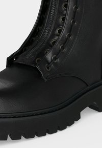 Botte de combat en cuir noir avec une semelle à crampons épaisse, des œillets métalliques, des lacets noirs et un détail de fermeture éclair central.