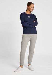 Sweatshirt bleu marine avec col rond et logo discret, associé à un pantalon de survêtement gris clair. Le modèle porte des baskets blanches, les bras croisés.