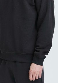 Felpa nera con collo rotondo, polsini e orlo ribattuti. Tessuto morbido e strutturato. Cuciture visibili lungo le cuciture. Design casual.