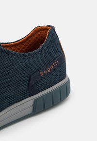 Primer plano de una zapatilla azul marino con forro interior naranja, parte superior de punto texturizado, suela gris y logo "bugatti" en el lateral.