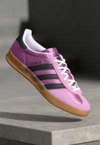 Lyserød ruskinds sportssko med sorte striber og hvide snørebånd. Har en glat tunge og en gummisål. Modelnavnet "Gazelle" er synligt.