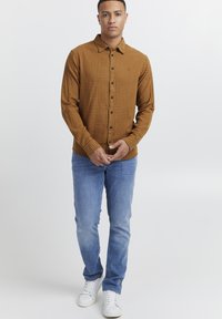 Camisa de cuadros marrón, de botones en la parte delantera y de mangas largas, combinada con jeans azules claros y zapatillas blancas. Fondo liso.
