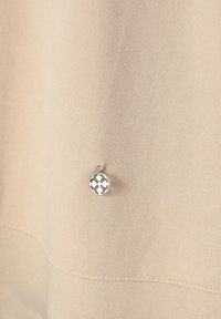 Béžová tkanina s hladkým povrchem, na které je malý kovový stříbrný přívěsek s vyřezávaným designem. Šití je viditelné blízko lemu.