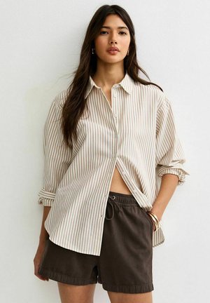 Jeune femme aux longs cheveux foncés portant une chemise à boutons beige surdimensionnée à rayures et un short marron foncé à cordon, debout contre un mur uni.
