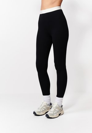 Osoba w czarnych legginsach z wysokim stanem, białych skarpetkach do kostek i jasnobeżowych butach sportowych, stojąca na tle gładkiej białej ściany.