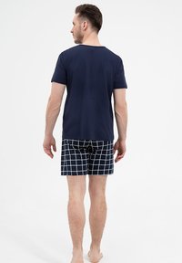 Granatowa koszulka z krótkim rękawem zestawiona z wzorzystymi szortami kąpielowymi w białą kratę. Widok z tyłu, model stoi boso.