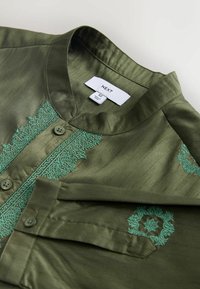 Olivgrünes Hemd mit Mandarinkragen, verziert mit türkisfarbenen Stickereien entlang der Knopfleiste und an der Brusttasche. Zwei Knöpfe sichtbar.
