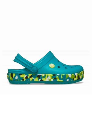 Zueco tipo slip-on color azul verdoso con orificios de ventilación y correa ajustable en el talón, con suela de patrón de camuflaje multicolor en verde, azul, blanco y negro.