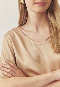 Beige Satinbluse mit V-Ausschnitt und kurzen Ärmeln. Glatte Textur, minimale Nähdetails, kombiniert mit goldenen Ohrringen.