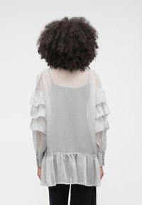 Harmaa läpikuultava paita, jossa on röyhelöiset hihat, röyhelöhelma ja napilliset kalvosimet. Pehmeä, kevyt kangas kerroksittaisella designilla.