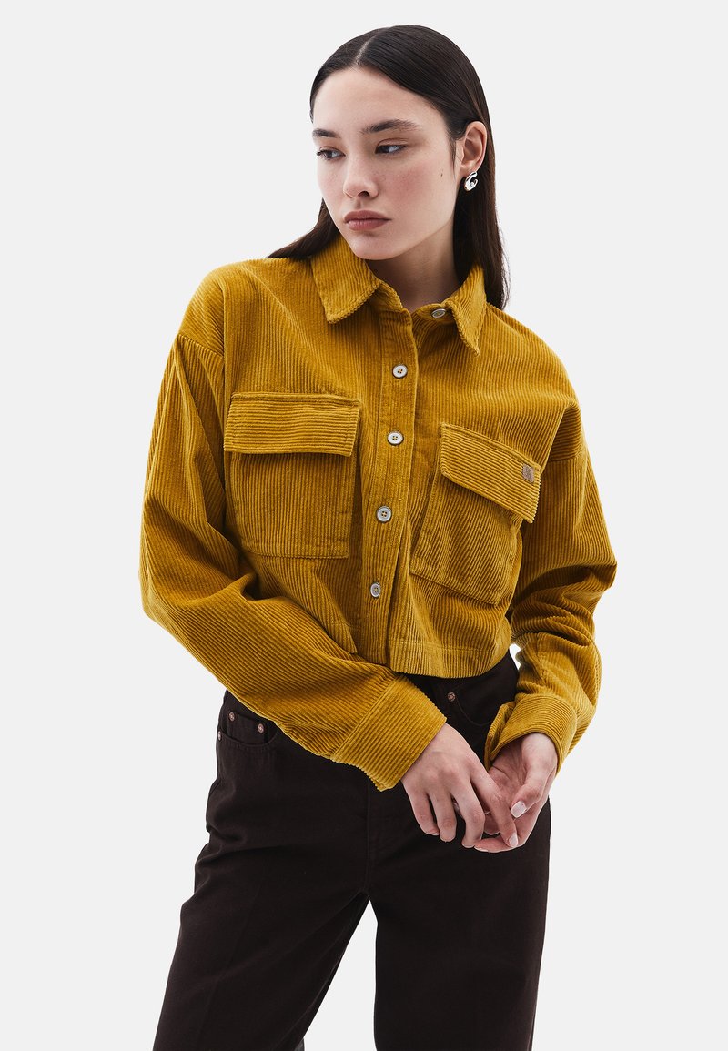 Hořčicově žlutá manšestrová košile s límcem, dvěma náprsními kapsami a knoflíky na přední straně; model má na sobě tmavě hnědé kalhoty.