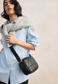 Sort quiltet crossbody-taske med gulddetaljer, kombineret med en blåstribet skjorte med knapper og et gråt uldtørklæde draperet over skuldrene.