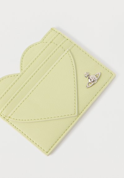 Λεπτή κίτρινη δερμάτινη θήκη καρτών με καπάκι σε σχήμα καρδιάς και μικρό ασημένιο λογότυπο με σφαιρίδιο σε λευκό φόντο.