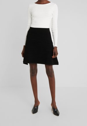 Frau trägt ein weißes, eng anliegendes Langarmshirt, einen schwarzen A-Linien-Rock und schwarze, spitz zulaufende High-Heel-Mules vor neutralem Hintergrund.