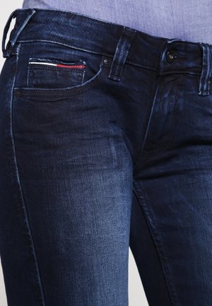 Vaqueros de mezclilla azul oscuro en una persona, mostrando los bolsillos delanteros, trabillas para cinturón y un pequeño detalle en rojo, blanco y azul en el bolsillo para monedas.