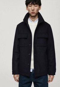 Svart ullblandad jacka med krage, två framfickor, knappstängning och mjuk textur, bärs över en vit tröja och en blå skjorta.