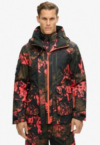 Chaqueta impermeable con una base negra y patrones abstractos en rojo y marrón, con cierre delantero, capucha ajustable y múltiples bolsillos.